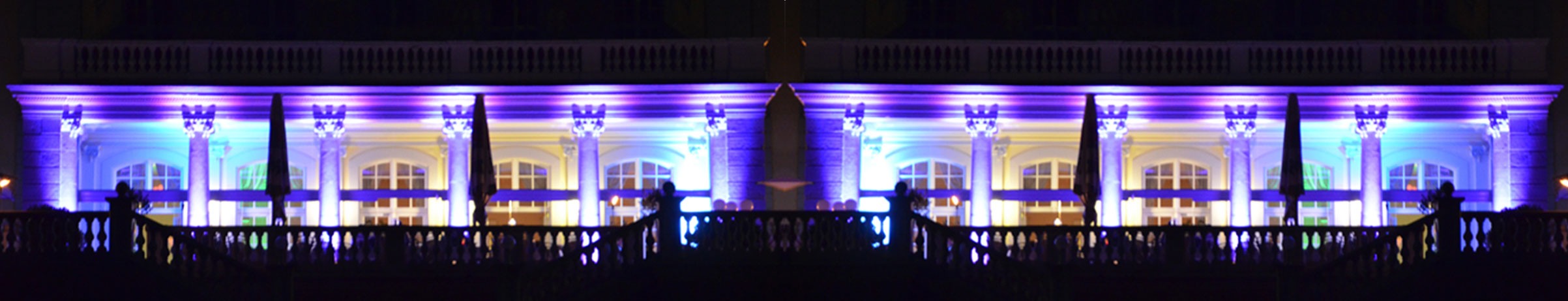 Schlosshotel von Außen bei Nacht, große Terasse mit 6 Säulen wird fantastisch in lila, türkis und gelb beleuchtet, Foto gespiegelt und nebeneinander